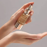 A person's hands holding a gold-toned cosmetic spray bottle labeled Cellulacore Cell-Lifting Serum, with the liquid being dispensed onto an open palm against a soft gray background.