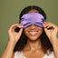 A woman with curly dark hair wearing a lavender-purple satin sleep mask, holding it in place with both hands against a solid olive green background.