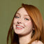 A close-up portrait of a smiling woman with long red hair, wearing gold hoop earrings and a gold sequined top against a muted olive green background.