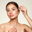 Woman applying a dropper of skincare product to her face against a plain background