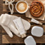 A styled flat-lay featuring Jane Rideau skincare products, including a tube of hand cream and a small lip balm tube, arranged with cream knit gloves, a cup of latte, a cinnamon roll, and silver accessories on a rustic wooden plank background.